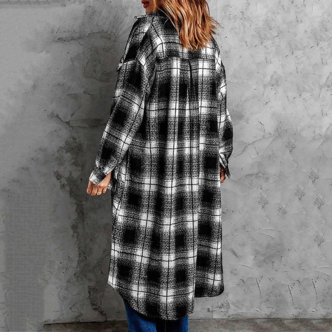 A person with wavy brown hair, seen from behind, layers a long black-and-white plaid button-down shirt jacket over blue jeans, standing against a textured gray concrete wall.