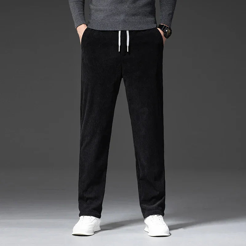 A person wearing a gray sweater, black men's smart casual pants with a zipper fly, and white sneakers stands with hands in pockets against a plain, dark background. Only the body from shoulders down is visible.