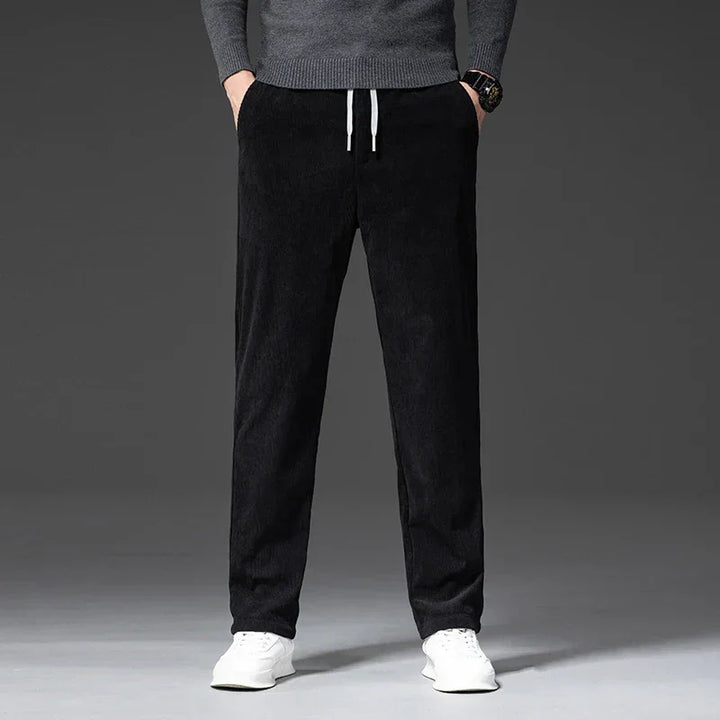 A person wearing a gray sweater, black men's smart casual pants with a zipper fly, and white sneakers stands with hands in pockets against a plain, dark background. Only the body from shoulders down is visible.