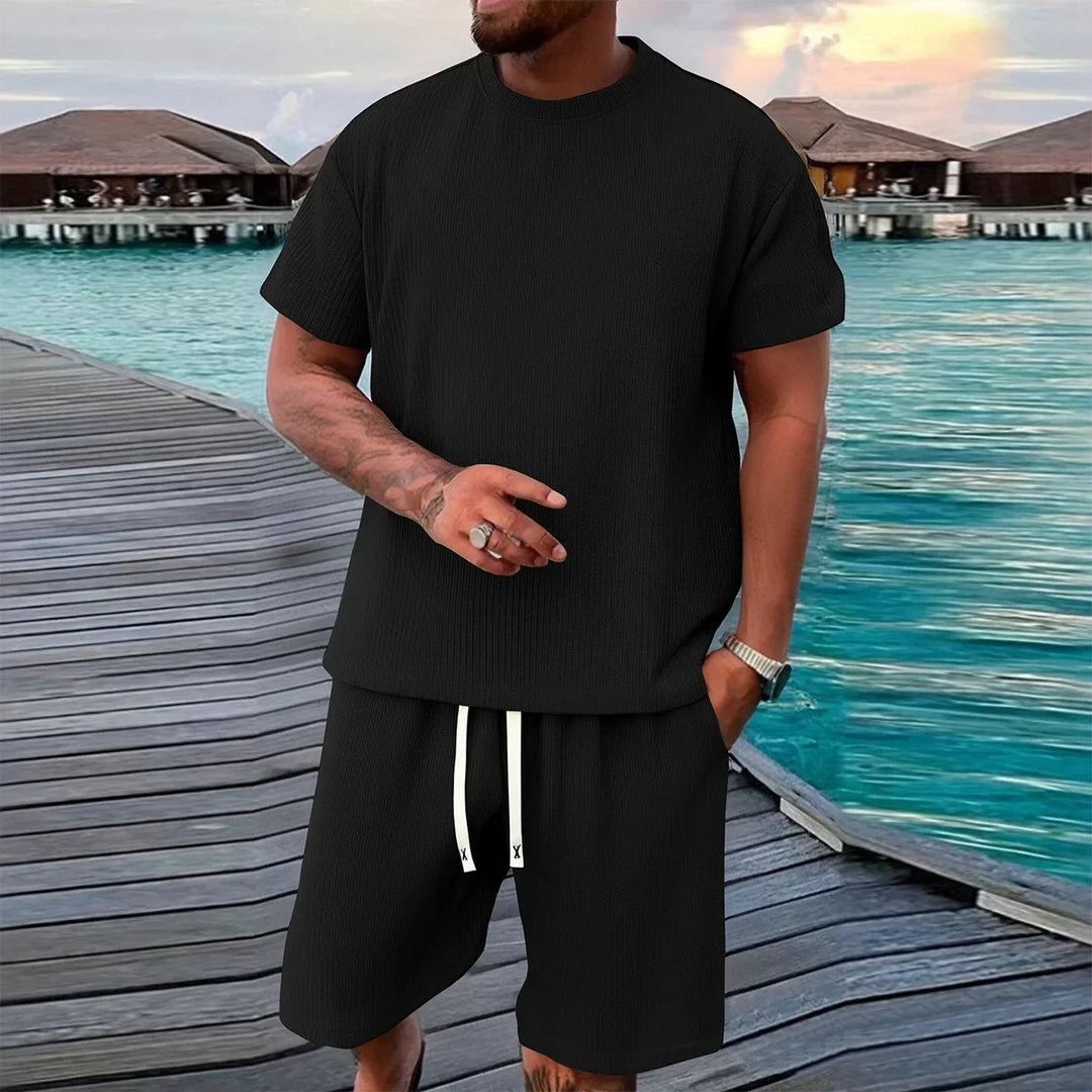 Man in black short sleeve shirt and shorts set standing on boardwalk by turquoise water, Rovelli fashion