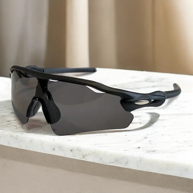 A pair of black, wraparound Polarized Cycling Sunglasses with dark lenses rests on a white marble surface. Beige curtains are visible in the blurred background.