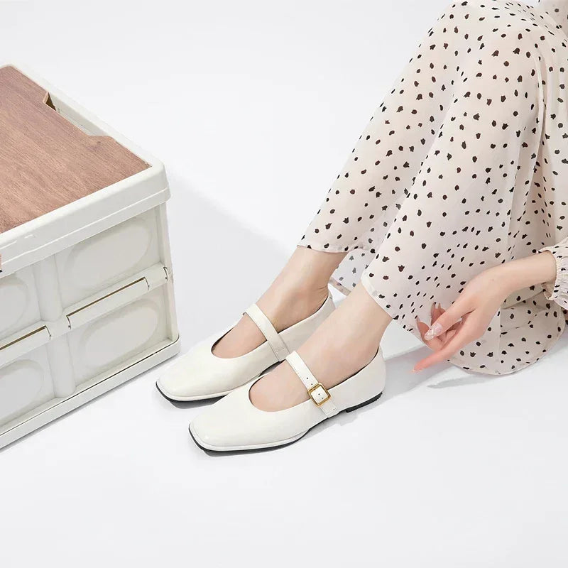 A person wearing cream-colored Augusta Leather Ballet Flats and cream pants with black polka dots sits on the floor next to a white and wooden storage box, with one hand resting on the ground.