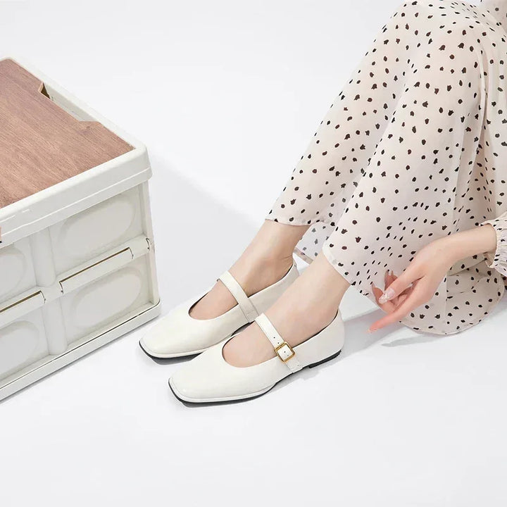 A person wearing cream-colored ballet flats and polka dot pants sits on the floor next to a white and wood storage box, extending their hand toward the ground.