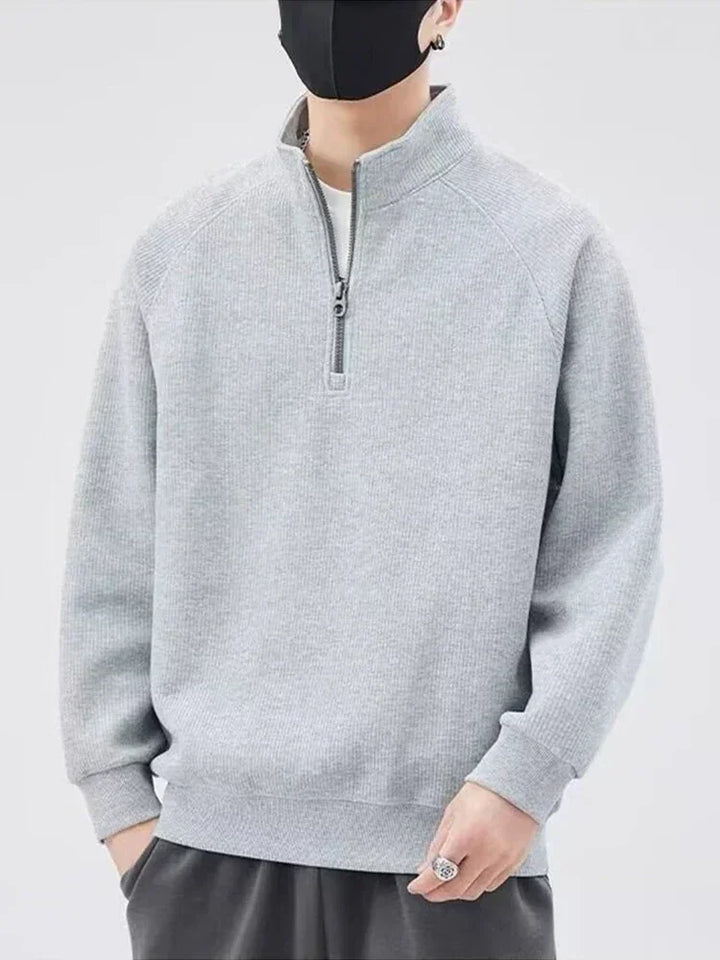 A person wearing a light gray zip-up sweater with a stand-up collar, dark pants, and a black face mask stands against a plain light background. Their hands are relaxed, and the face is mostly covered.