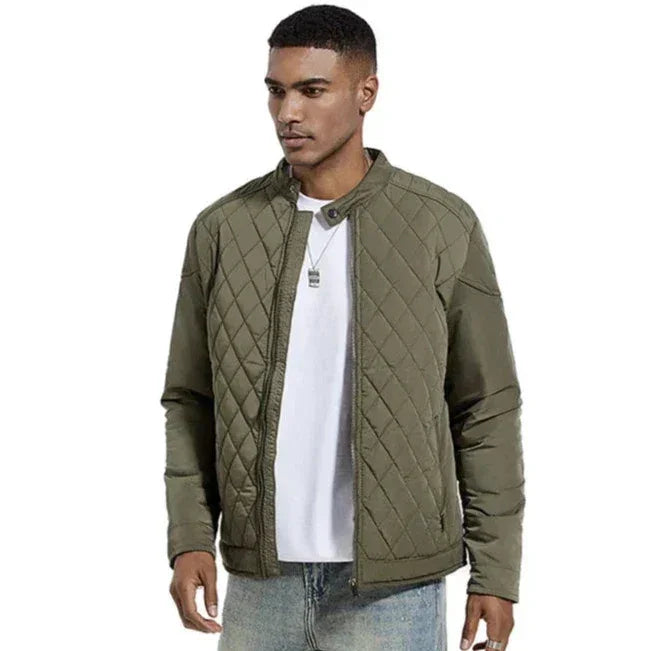 A man wearing a wind-resistant quilted olive green jacket over a white t-shirt and light blue jeans stands with his hands relaxed at his sides, looking slightly to the left.