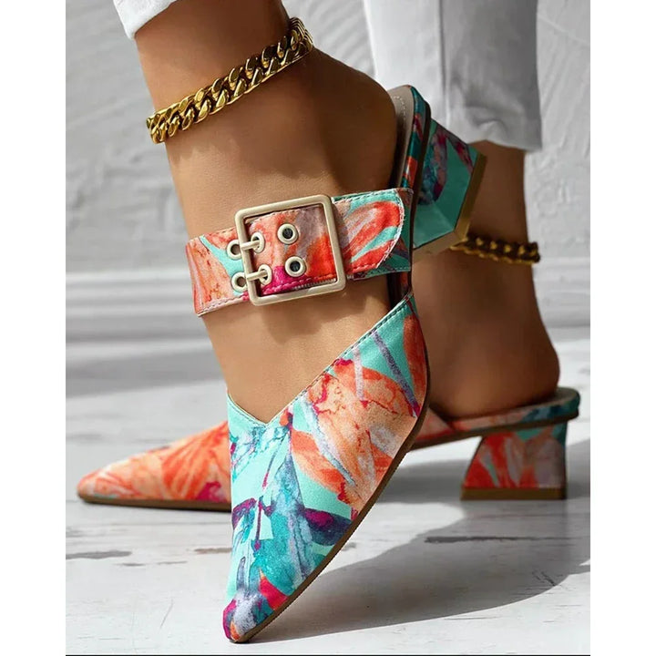 A close-up of a foot wearing a pointed-toe, floral-print mule heel with a bold square buckle and gold anklet; these vibrant summer shoes in blue, orange, and coral are paired stylishly with white pants.