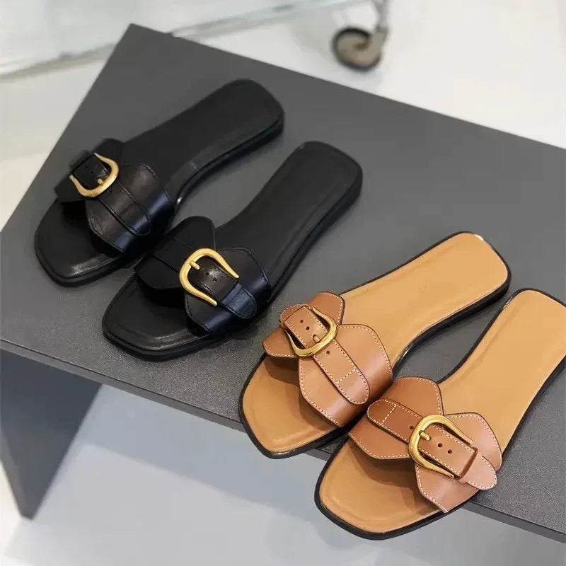 Two pairs of slip-on leather slides with large gold buckles—one in black and the other in tan with a beige insole—are displayed on a gray surface.