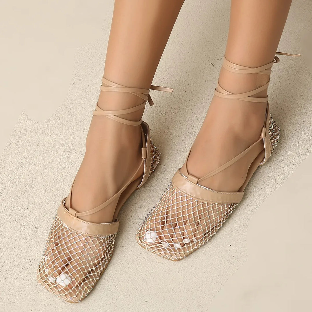 A pair of feet wearing beige, mesh-patterned flats with pointed toes and nude ankle wrap straps, a true summer essential shown against a light, neutral background.