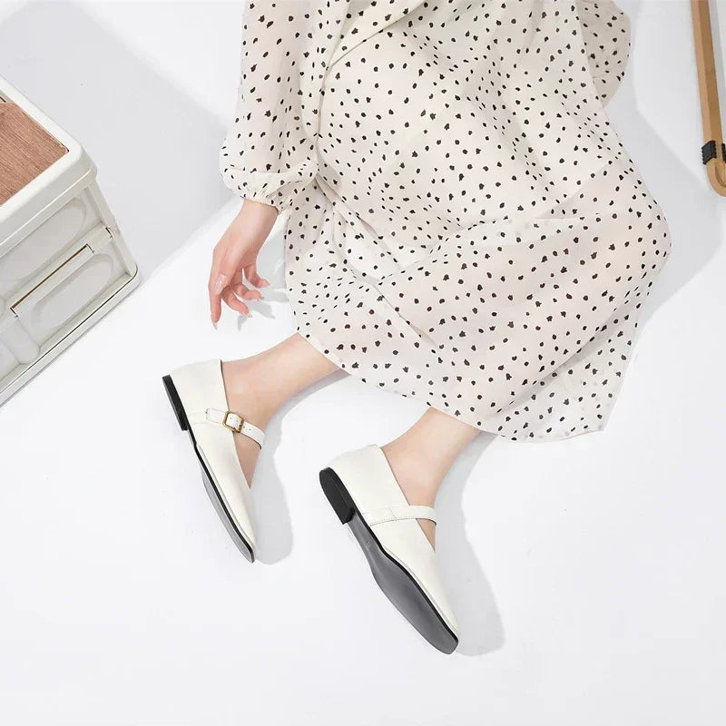 A person wearing a white dress with black polka dots sits on the floor, showing off handmade leather flats with a buckle detail. The scene is bright with minimalist white decor.