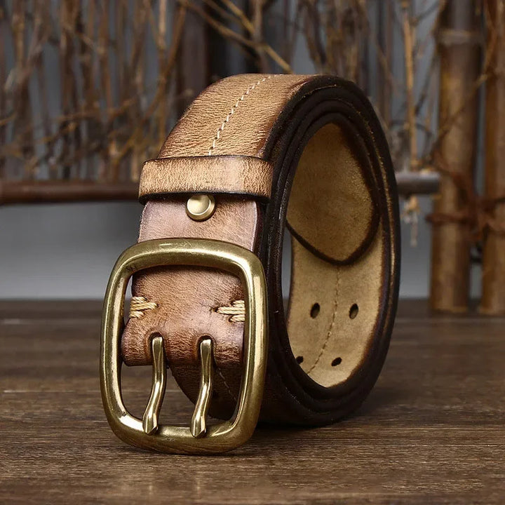 A thick, genuine leather belt with a copper buckle is coiled on a wooden surface, set against a rustic backdrop of wooden branches and a gray wall—showcasing true heritage craftsmanship.