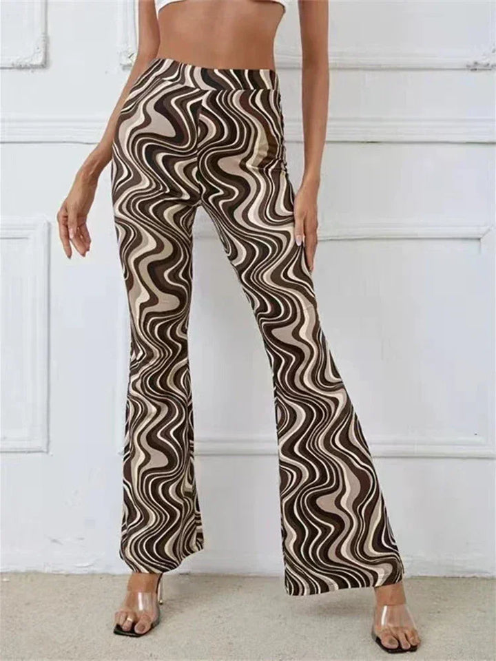 A person wearing trendy statement pants with a brown, beige, and black wavy swirl pattern and clear-heeled sandals stands against a white wall. Only the lower torso and legs in these flared pants are visible.