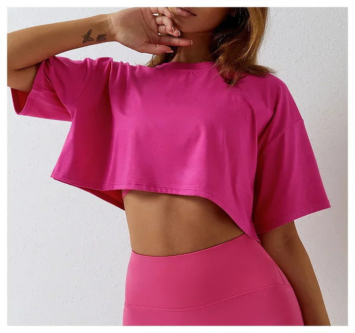 A woman wearing a bright pink, short-sleeve cotton crop top and matching high-rise pants stands against a white background, with one hand touching her chin. A small tattoo is visible on her inner arm.