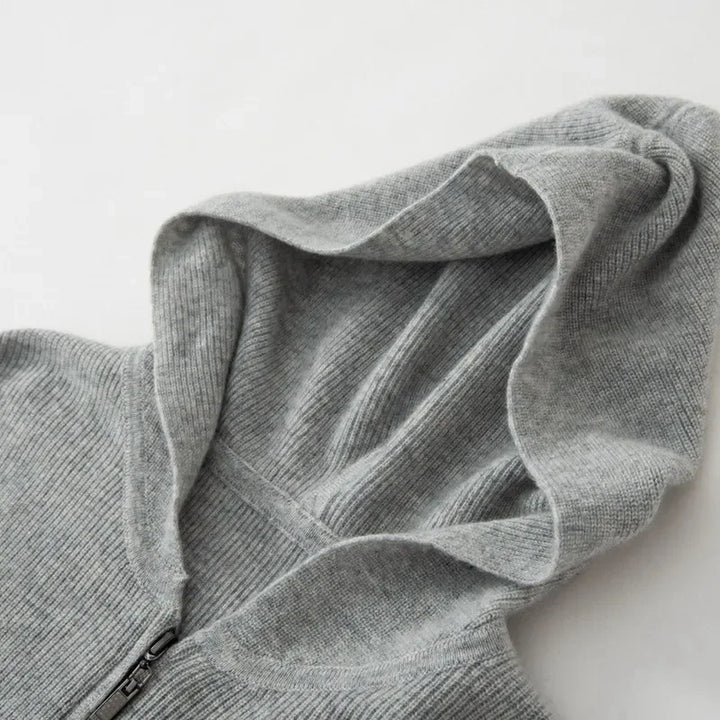 A close-up of a light gray, ribbed knit cashmere hoodie with a zipper, showing the luxurious cashmere texture and lightweight insulating fabric, with the hood laid flat on a white surface.