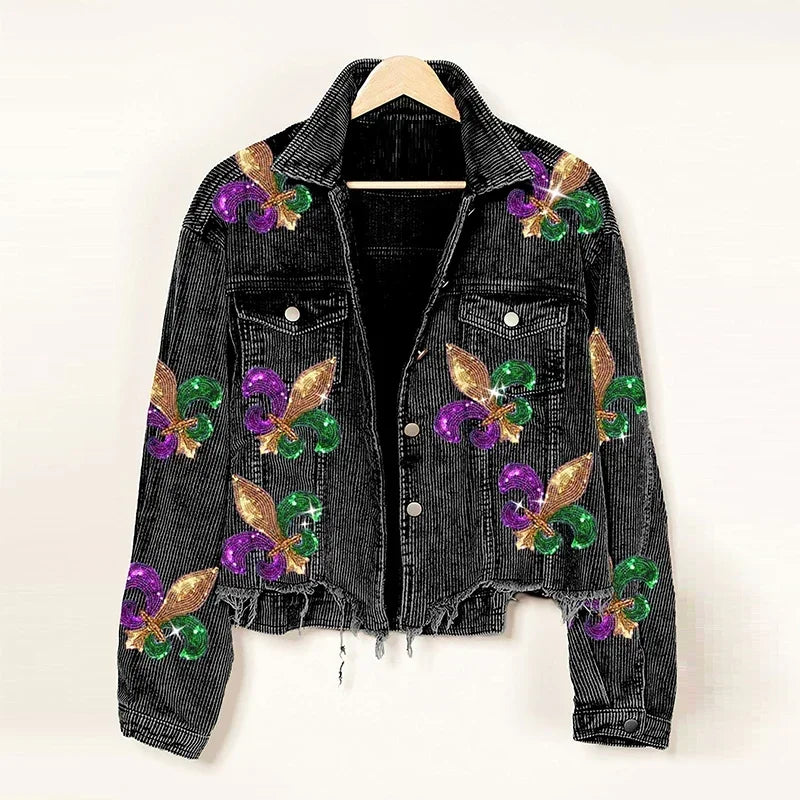 A black denim jacket with a cropped relaxed fit, decorated with glittery gold, green, and purple fleur-de-lis designs, hangs on a wooden hanger against a plain background.