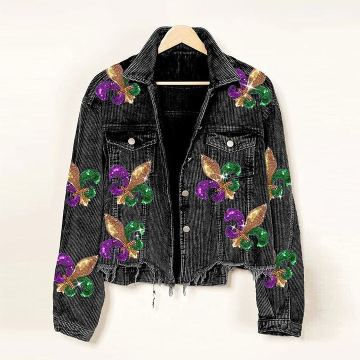 A black denim jacket with a cropped relaxed fit, decorated with glittery gold, green, and purple fleur-de-lis designs, hangs on a wooden hanger against a plain background.