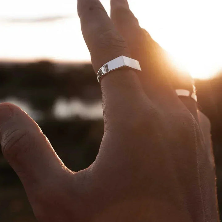 A close-up of a hand with two rings, one stainless steel and one black, raised against a bright sunset background. The sunlight highlights the scratch-resistant rings and casts a warm glow on the skin.