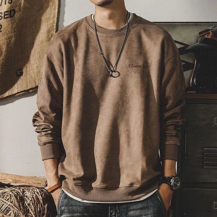 A man wears a relaxed fit crewneck in a loose brown, earth-toned style with a round neckline, black necklace with a ring pendant, and watch. His face isn’t visible as he stands casually with hands in his pockets.