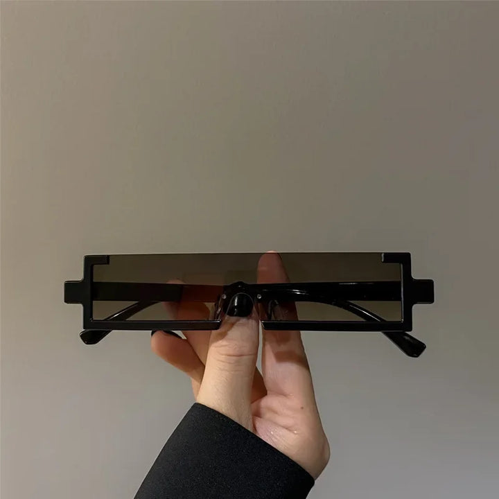 A hand is holding a pair of rectangular, geometric sunglasses with thick black frames and dark lenses that offer UV400 protection against a plain gray background.