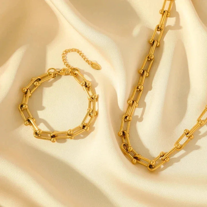 A gold bracelet and matching 316L stainless steel necklace with bead accents are displayed on soft, cream-colored fabric, highlighted by gentle natural light.