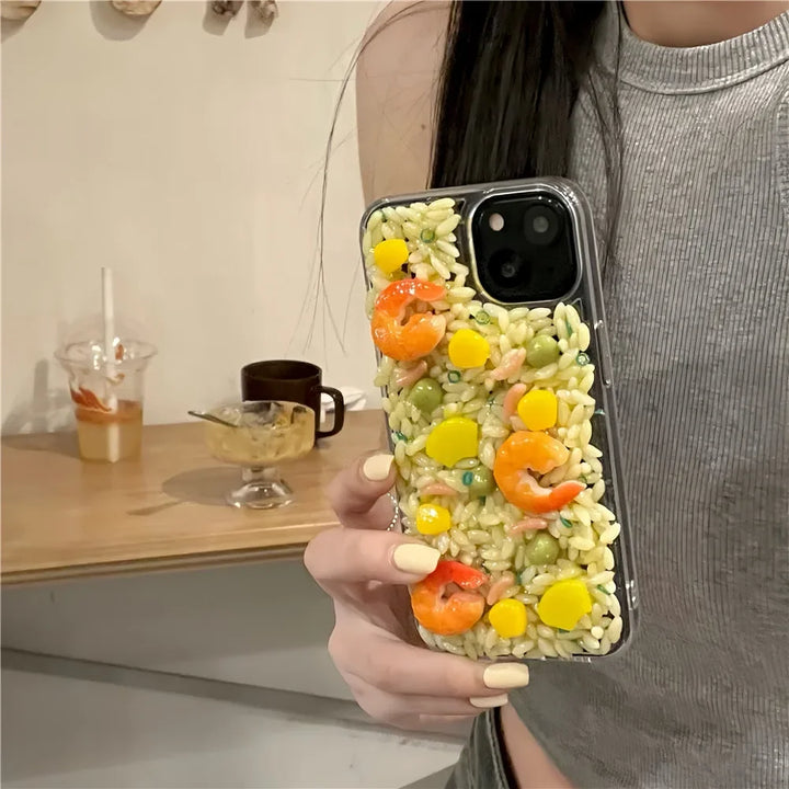 A person holds a phone with a shock-absorbent case decorated to look like fried rice with shrimp, corn, peas, and yellow pieces. The person has pale yellow nails and wears a gray sleeveless top. A table with cups is blurred in the background.
