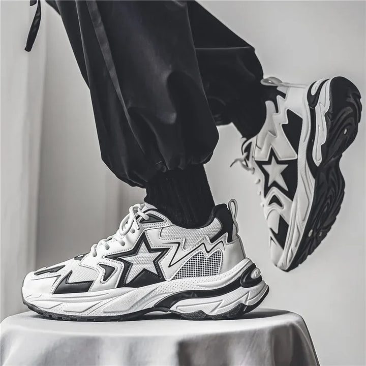 A person wearing black pants and black socks stands on a stool, showcasing chunky white sneakers with bold black star designs, a breathable mesh upper, and striking patterns on the sides and soles.