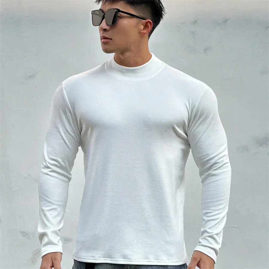 A man wearing dark sunglasses and a fitted white long-sleeve shirt with a mock neck collar stands against a plain light gray background, looking to the side with a confident posture.