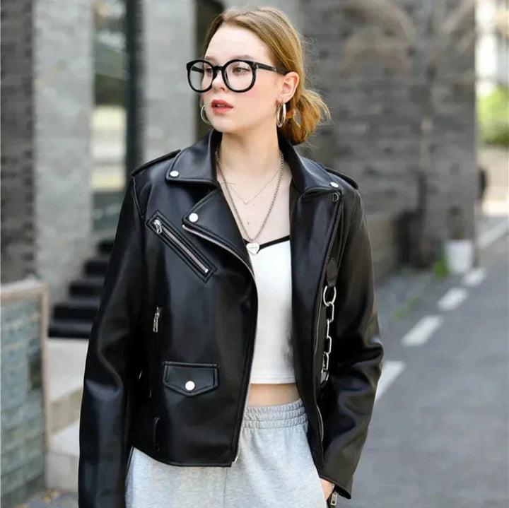 A woman wearing black-rimmed glasses, a black leather jacket, a white crop top, and gray sweatpants with an adjustable waist belt stands outdoors on a city street, looking to the side.