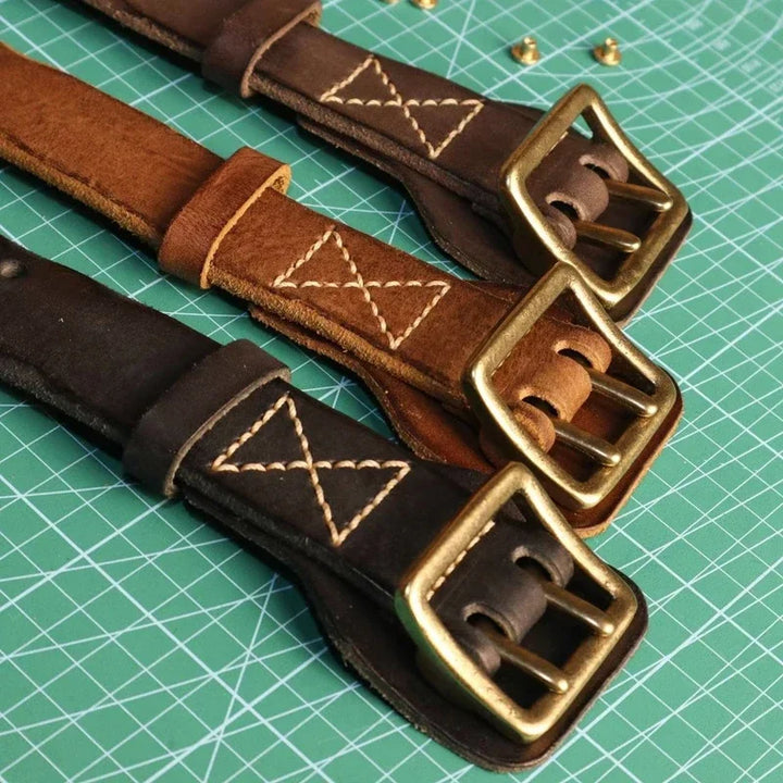 Three handmade cowhide belts in dark brown, brown, and black with brass buckles are laid out on a green cutting mat. Each features sturdy stitching in a diamond and X pattern near the buckle, offering a rugged aesthetic.