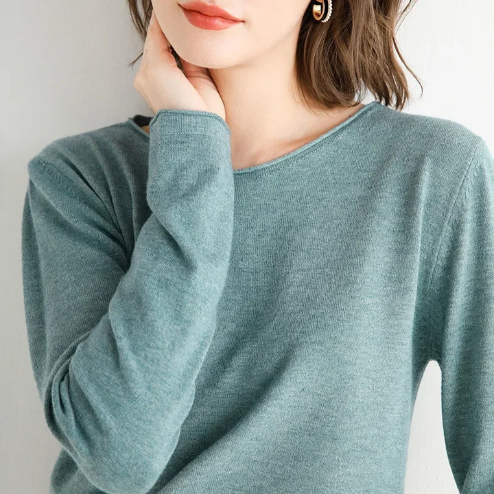 A woman in a relaxed fit, light teal long-sleeve top rests her chin on her hand. She has short brown hair, peach lipstick, and wears a gold earring. Her face is partly out of frame.