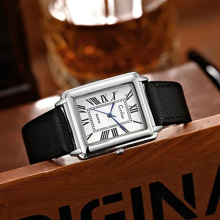 A rectangular luxury watch with a white dial, Roman numerals, blue hands, and a premium leather band rests on a wooden surface, with a blurry glass in the background.