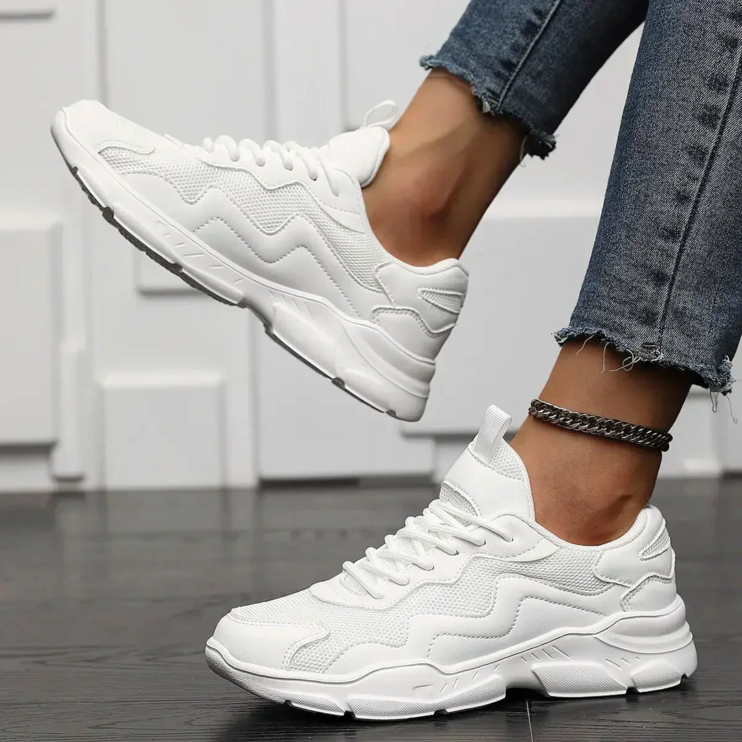 A person wearing white slip-on street shoes with frayed blue jeans sits with legs crossed, showing the sneakers’ side profile—lightweight mesh stands out against a white textured wall and dark wooden floor.