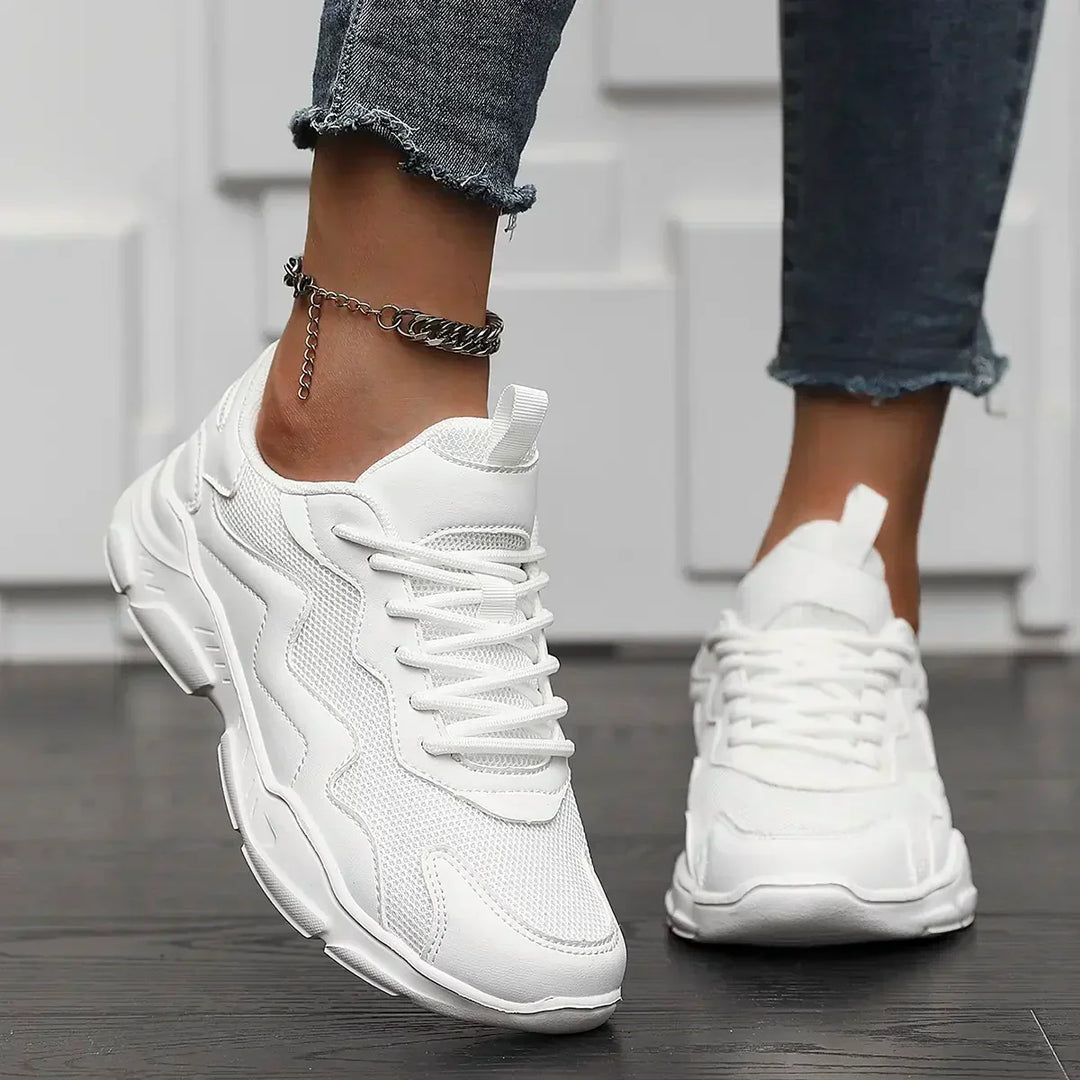 A person wearing white chunky sneakers with advanced shock absorption and ankle-length distressed jeans stands on a dark wooden floor, with one foot slightly forward, showcasing the shoes and a silver anklet.