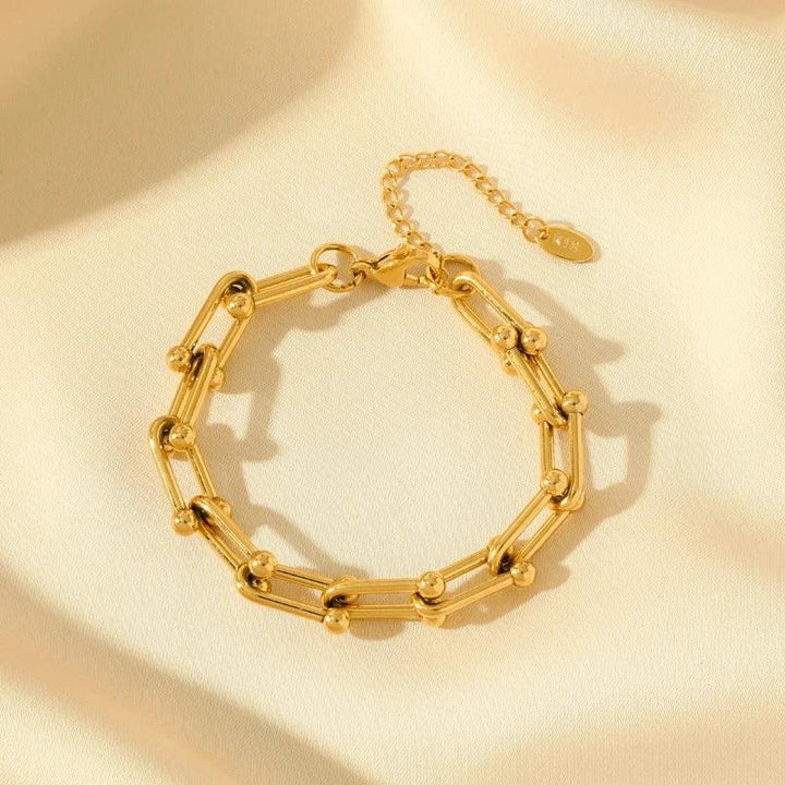 A gold statement chain bracelet with cylindrical links and small round accents, crafted from 316L stainless steel. Displayed on soft cream fabric, it features an adjustable extension chain and a small round tag.