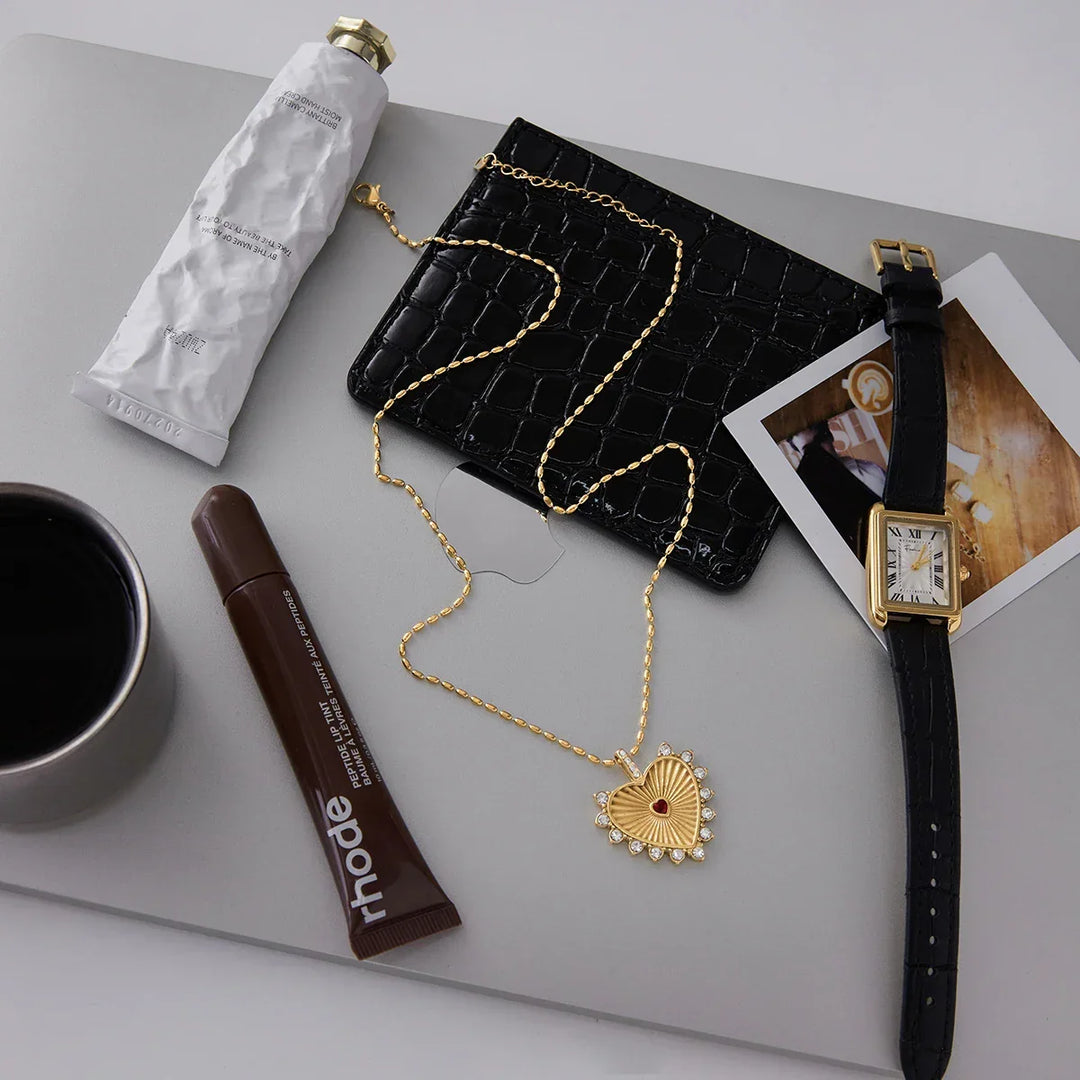 Flat lay of Rovelli gold heart necklace, black leather wallet, watch, and accessories on laptop