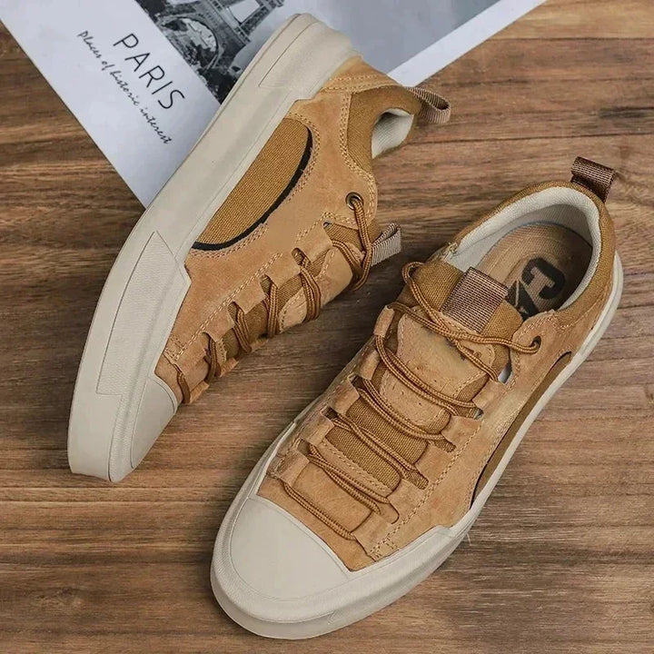 A pair of tan suede canvas shoes with beige durable rubber outsoles and brown laces displayed on a wooden surface, with a partial image of a Paris-themed magazine in the background.