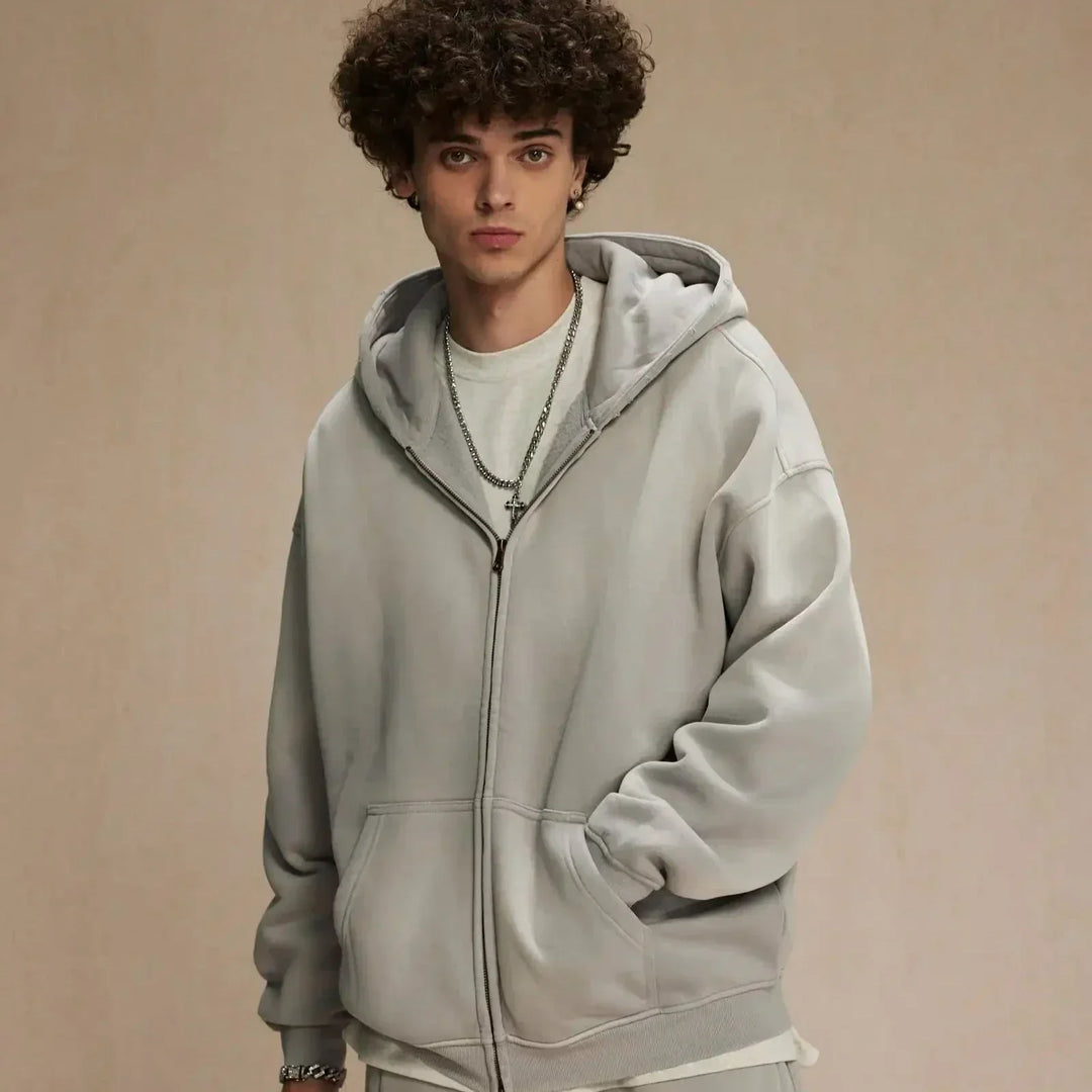 A young man with curly brown hair wears a relaxed fit, light gray zip-up hoodie over a white T-shirt. He stands against a plain beige background, looking at the camera with his hands in his pockets.