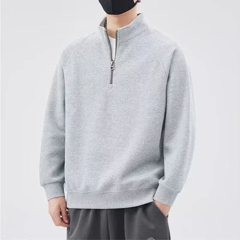 A person wearing a light gray, long-sleeve sweatshirt with a stand-up collar and half-zip closure, paired with dark pants, stands against a plain background. The relaxed fit outfit is complemented by a black face mask. Their face is mostly out of frame.