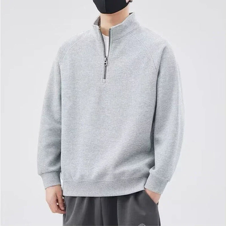 A person wearing a light gray, long-sleeve sweatshirt with a stand-up collar and half-zip closure, paired with dark pants, stands against a plain background. The relaxed fit outfit is complemented by a black face mask. Their face is mostly out of frame.
