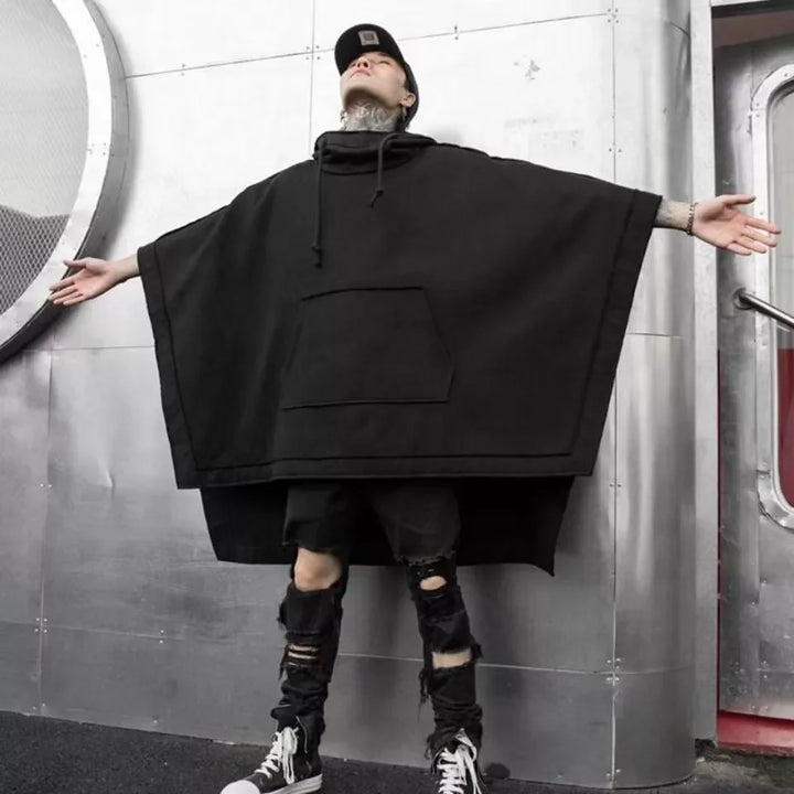 A person in a black streetwear poncho with a front pocket and ripped black jeans stands with arms outstretched in front of a metallic wall, looking upward. They wear a cap and black high-top sneakers.