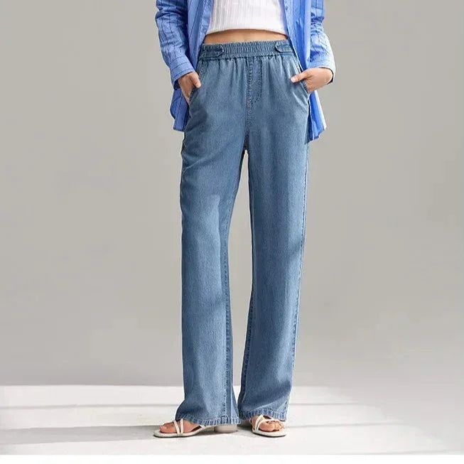 A person wearing wide-leg, light blue breathable denim pants with an elastic waistband, a white top, a blue shirt, and white sandals stands with hands in pockets against a plain background.