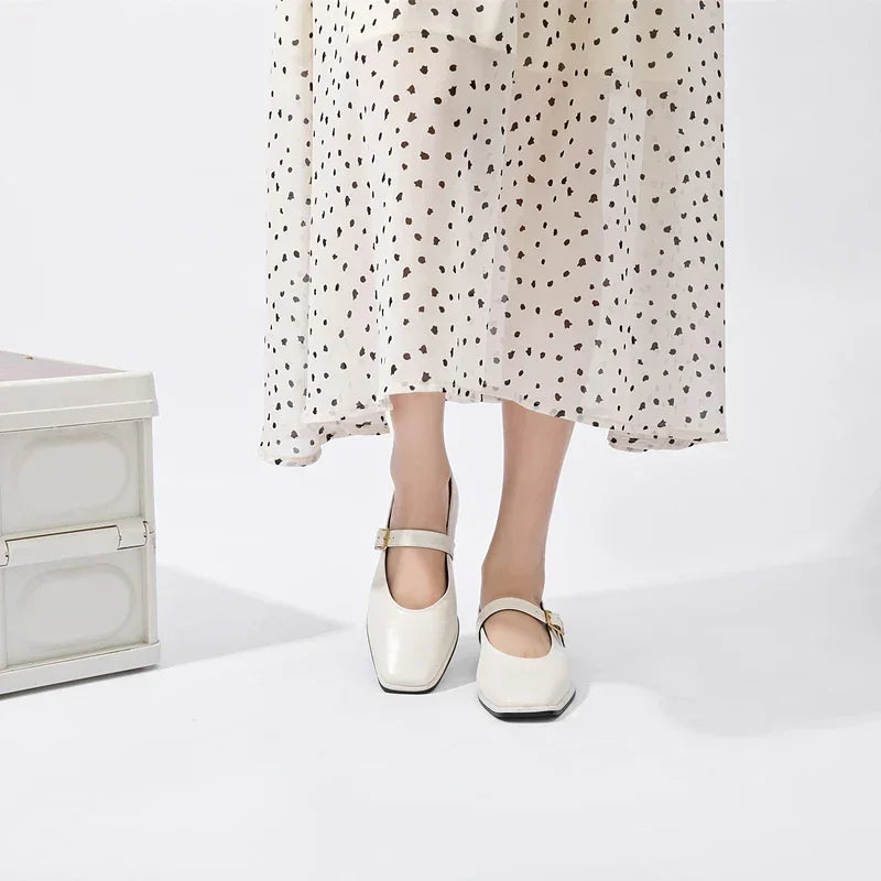A person wearing a white, ankle-length dress with black polka dots and classic white Mary Jane shoes stands next to a white trunk against a plain background.