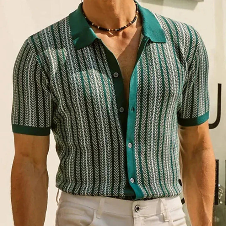 A man wearing a green patterned short-sleeve shirt with white arrow-like designs in a jacquard herringbone pattern, an open collar, blue buttons, white pants, and a black beaded necklace. His head is mostly out of the frame.