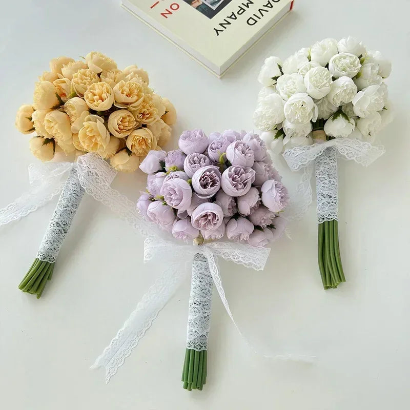 Three small bouquets of roses—yellow, light purple, and white—made with maintenance-free faux petals are wrapped in white lace and arranged on a white surface beside an open book.