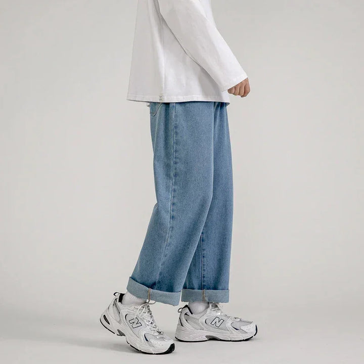 A person stands against a plain background wearing a white long-sleeve shirt, relaxed fit jeans in light blue with cuffs, white socks, and white New Balance sneakers. The persons head is not visible.