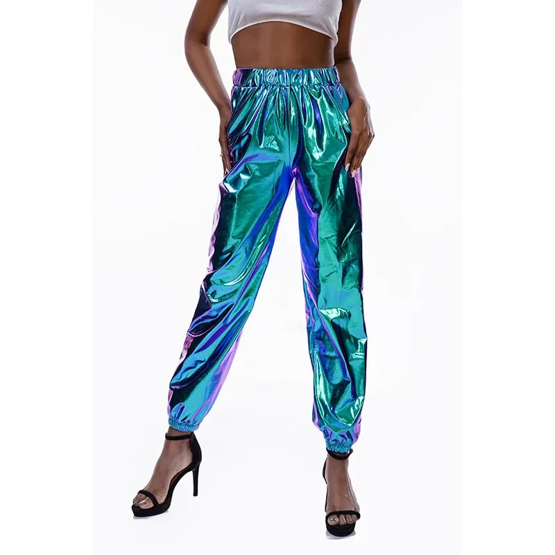 A person wearing shiny, iridescent turquoise jogger pants with an elastic high waist and holographic finish, paired with a white crop top and black high-heeled sandals, standing against a white background.