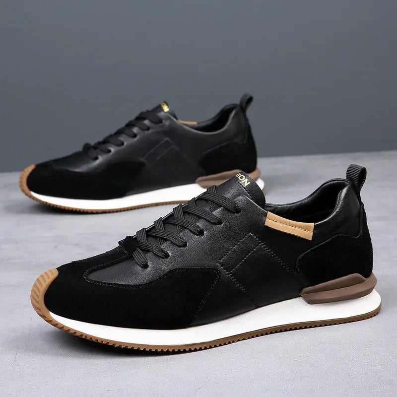 A pair of stylish black sneakers with white midsoles, brown accents on the heel and tongue, and black laces—crafted with top-grain leather—displayed on a gray surface with a plain gray background.