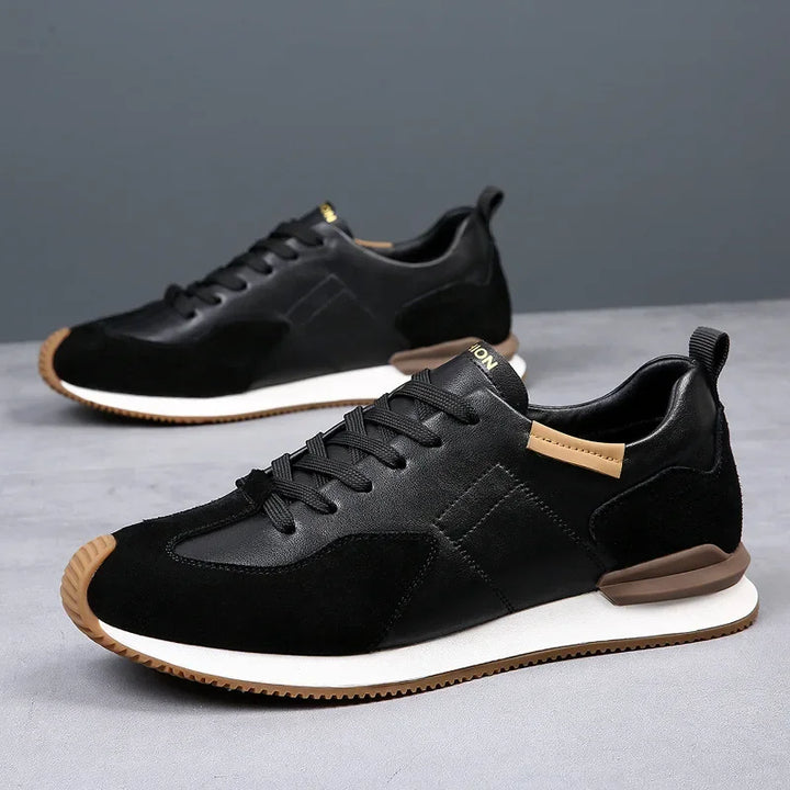 A pair of stylish black sneakers with white midsoles, brown accents on the heel and tongue, and black laces—crafted with top-grain leather—displayed on a gray surface with a plain gray background.