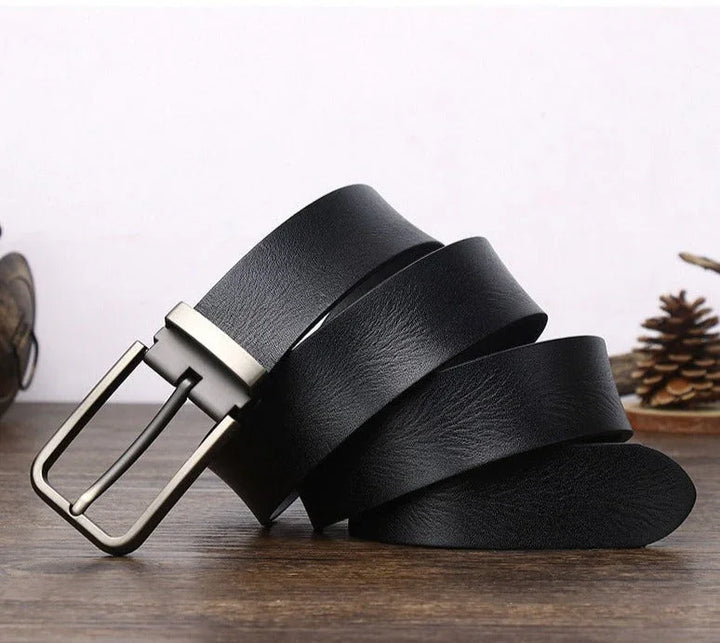 A black cowhide belt with a silver metal buckle is coiled on a wooden surface, with a pinecone and small decorative items blurred in the background.