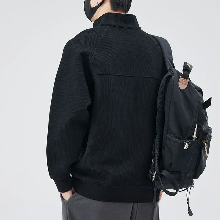 A person wearing a black jacket with a stand-up collar, gray pants, and a black face mask is seen from behind, carrying a black backpack over one shoulder against a plain light background.