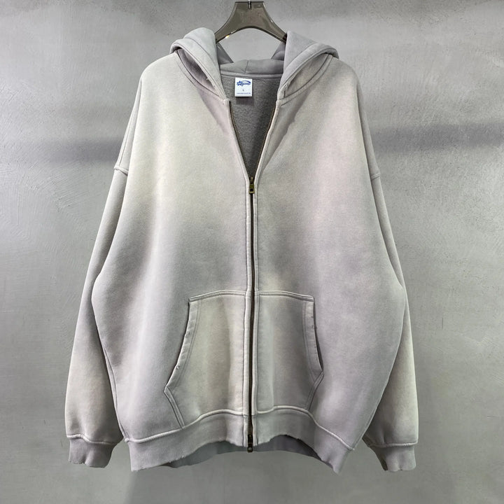 A light gray oversized zip-up hoodie with a relaxed fit, hood, and front pockets hangs on a clear hanger against a plain gray textured wall.